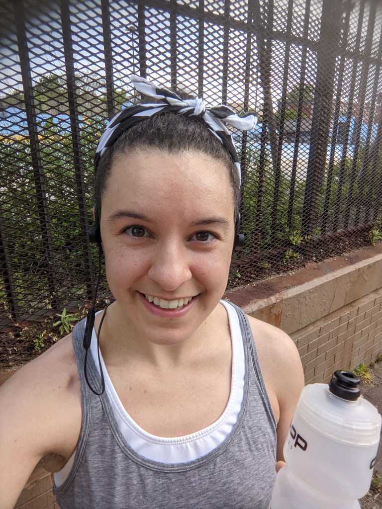 Shea - Smiling, but tired holding a water bottle and wearing workout gear.